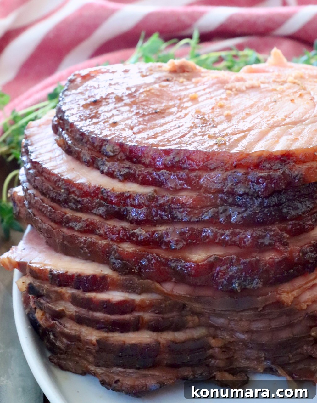 How to Smoke a Ham: Glaze, Time, and Temperature 2 Sliced spiral ham glazed to a shiny brown, stacked on a white plate. Fresh herbs in the background and a red-striped cloth