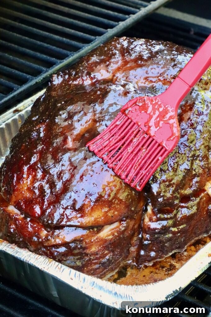 How to Smoke a Ham: Glaze, Time, and Temperature 9 A smoked ham in a foil tray is being glazed with sauce using a red brush, on a smoker.