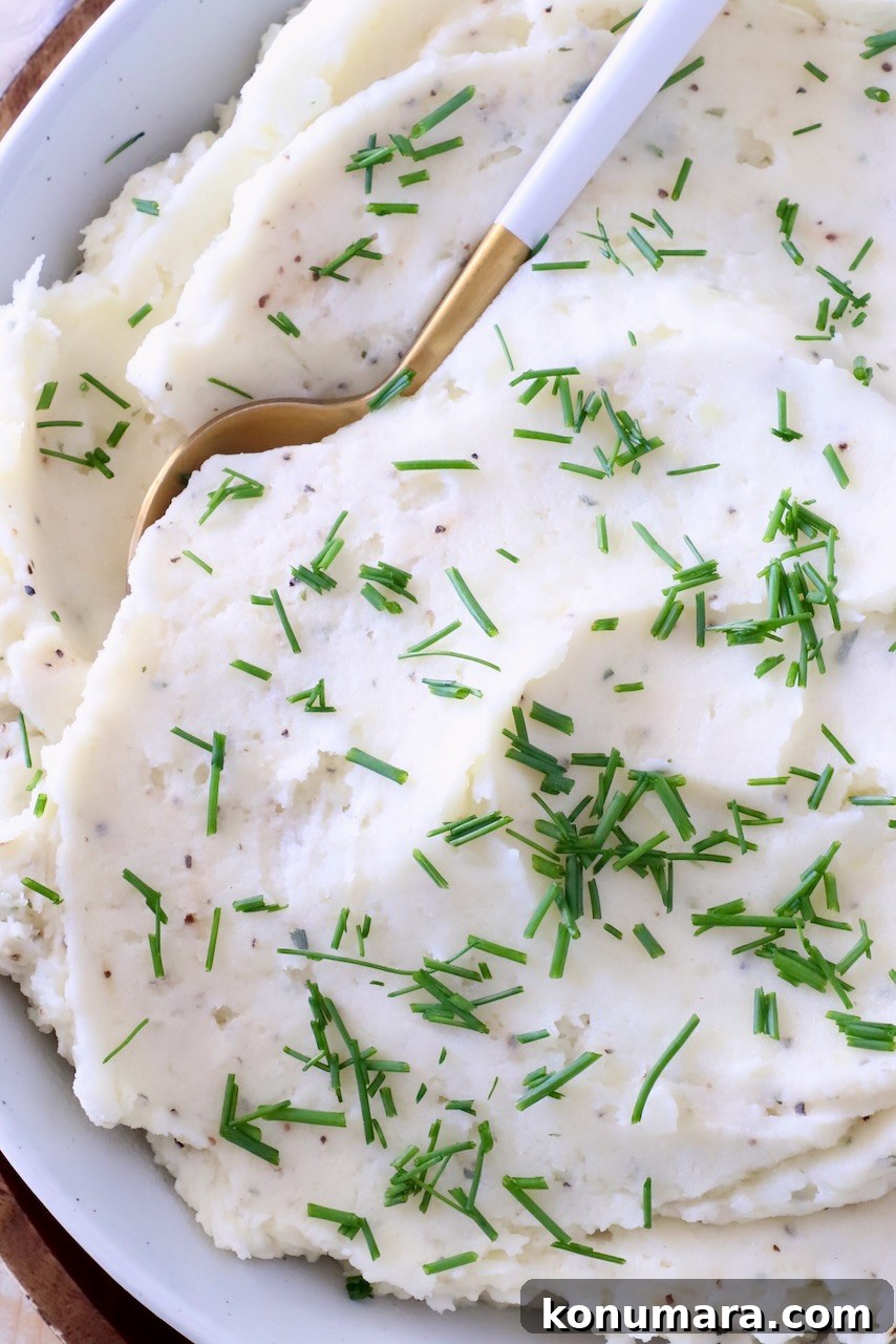 Creamy mashed potatoes garnished with fresh chopped chives in a white bowl. A gold and white spoon is dipped in the potatoes.