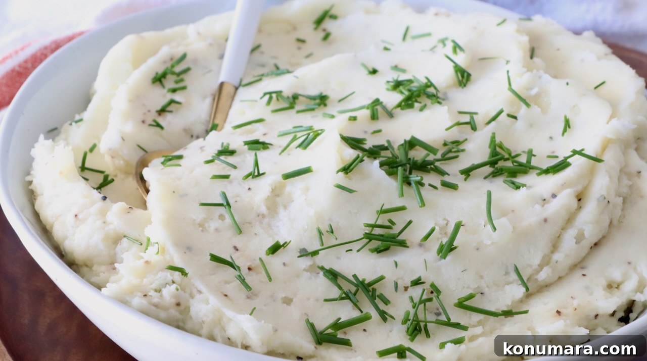 A white bowl filled with creamy mashed potatoes garnished with fresh green chives. A golden spoon rests in the dish.