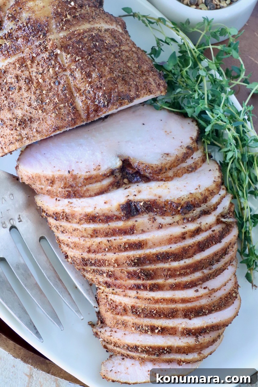 Sliced smoked turkey breast on a white platter, seasoned with spices. Fresh herbs are placed beside it, along with a serving fork.