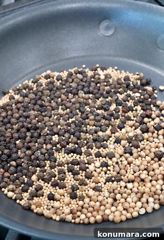 Assorted whole spices, including black peppercorns, mustard seeds, and coriander seeds, are being dry roasted in a dark frying pan.