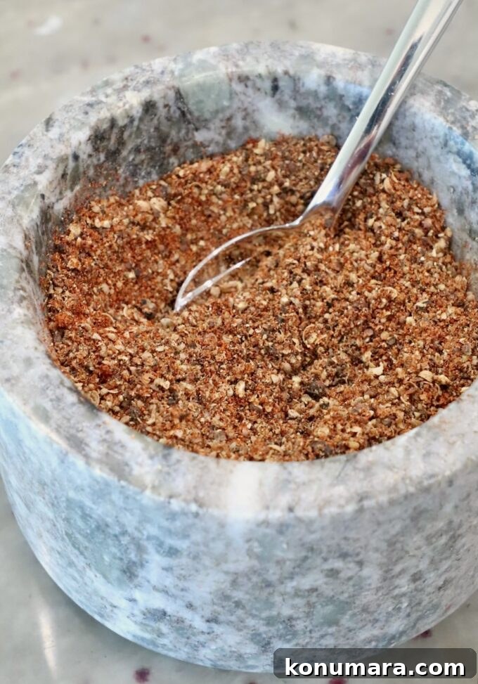 A close-up of a marble mortar filled with a coarse, mixed spice blend. A shiny metal spoon is partially submerged in the spices
