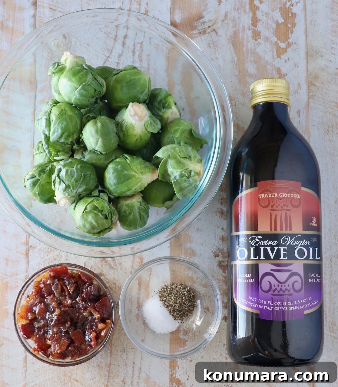 A bowl of Brussels sprouts, a bottle of olive oil, a small dish of bacon jam, and a bowl with salt and pepper are arranged on a wooden surface.