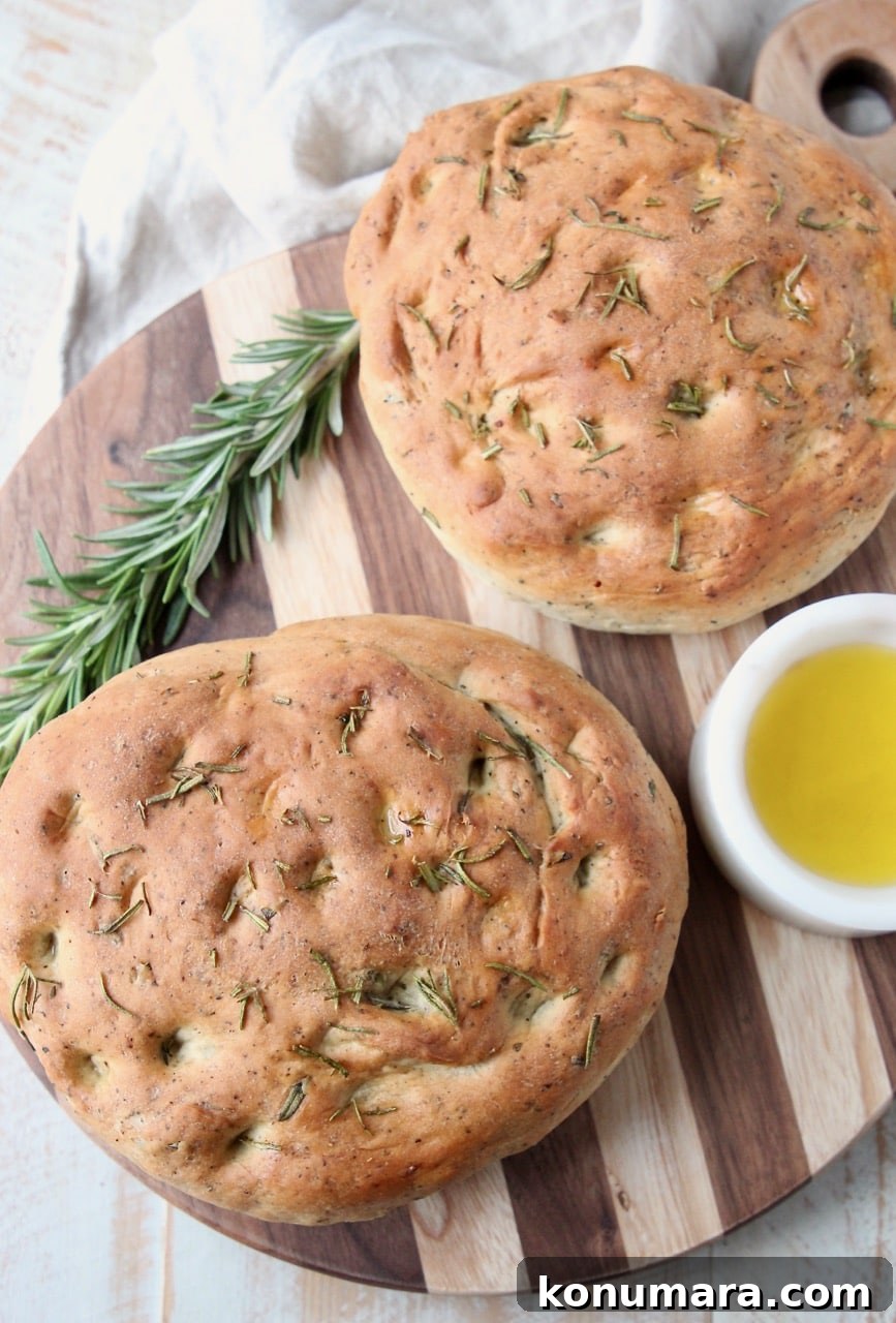 Two loaves of rosemary focaccia bread on wood cutting board with fresh sprig of rosemary and small white bowl of olive oil