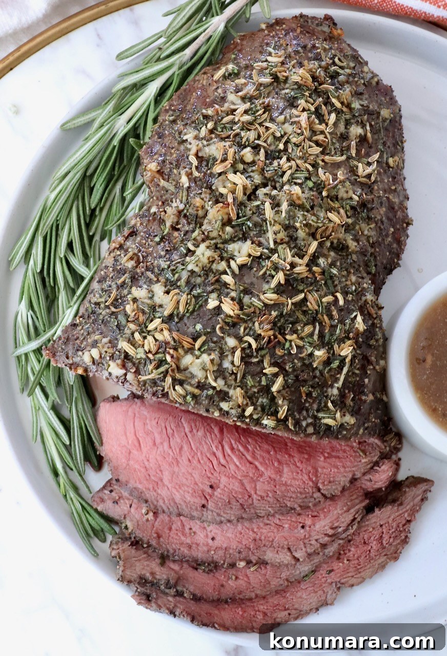 Sirloin Tip Roast Recipe for Tender, Juicy Results 9 Roasted beef topped with herbs, sliced on a white plate with a sprig of rosemary. A small bowl of gravy sits on the side.