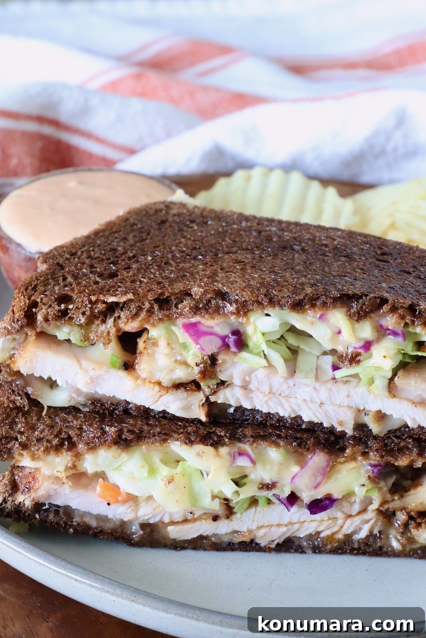Close-up of a Rachel sandwich with dark rye bread filled with sliced turkey, creamy slaw, and sauce. Served on a plate with wavy potato chips and a dipping sauce.