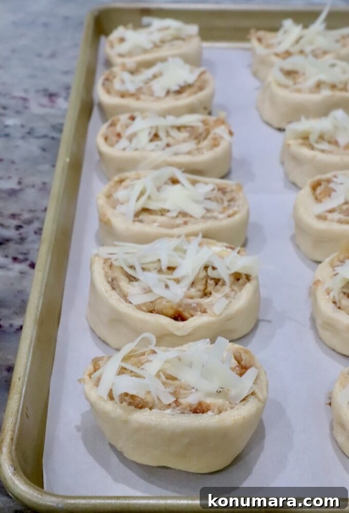 Close-up of unbaked caramelized onion rolls on a parchment-lined baking sheet. Each roll is topped with shredded cheese.