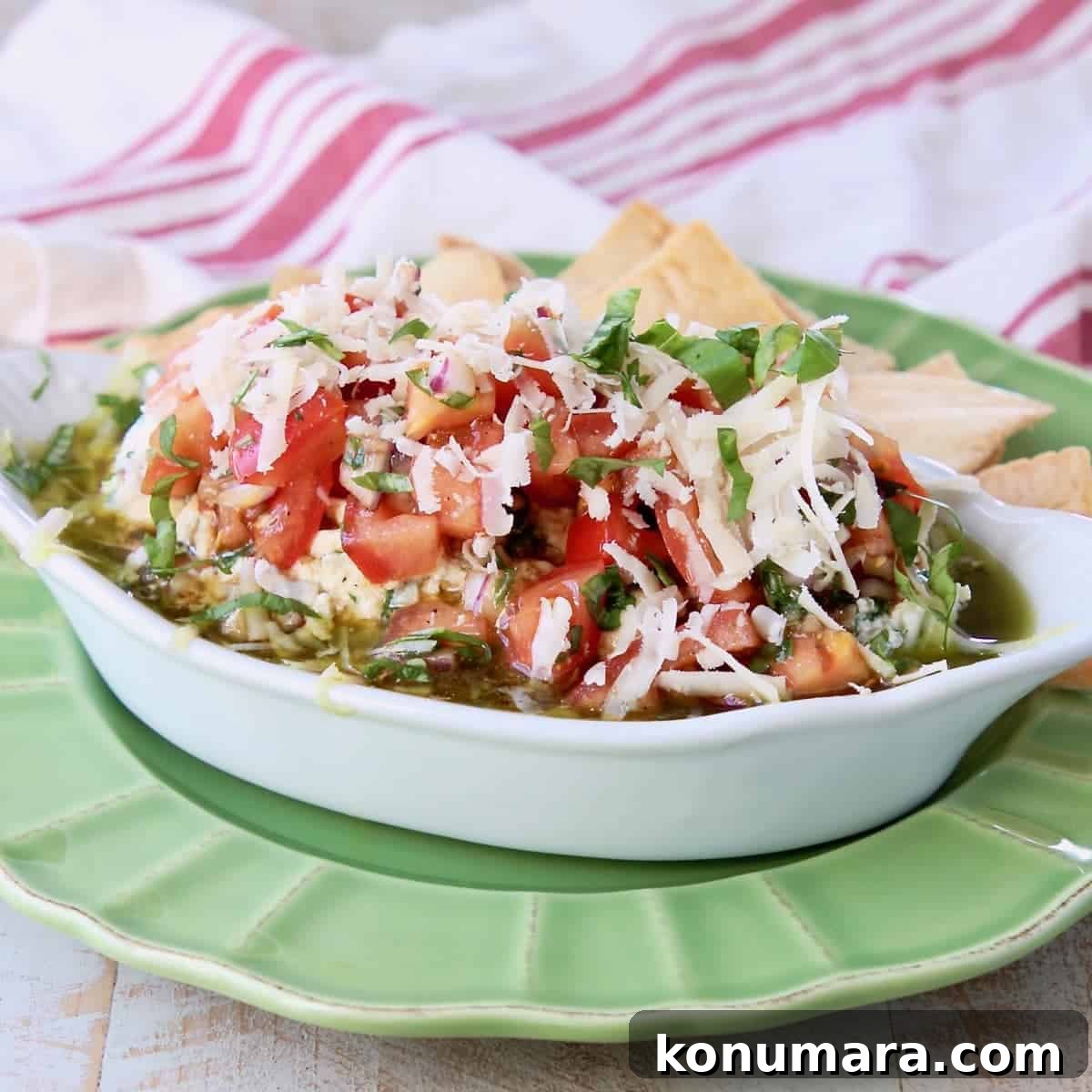 Italian 5 layer dip topped with tomato basil bruschetta in a white serving bowl sitting on a green plate with pita chips