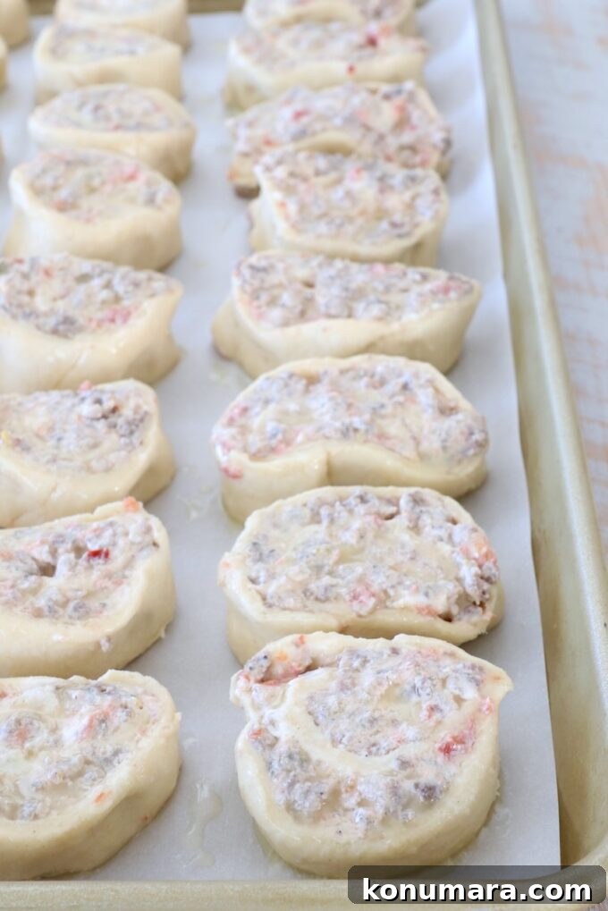 Rows of uncooked pastry pinwheels filled with a creamy mixture of sausage and tomatoes are neatly arranged on a parchment-lined baking sheet.
