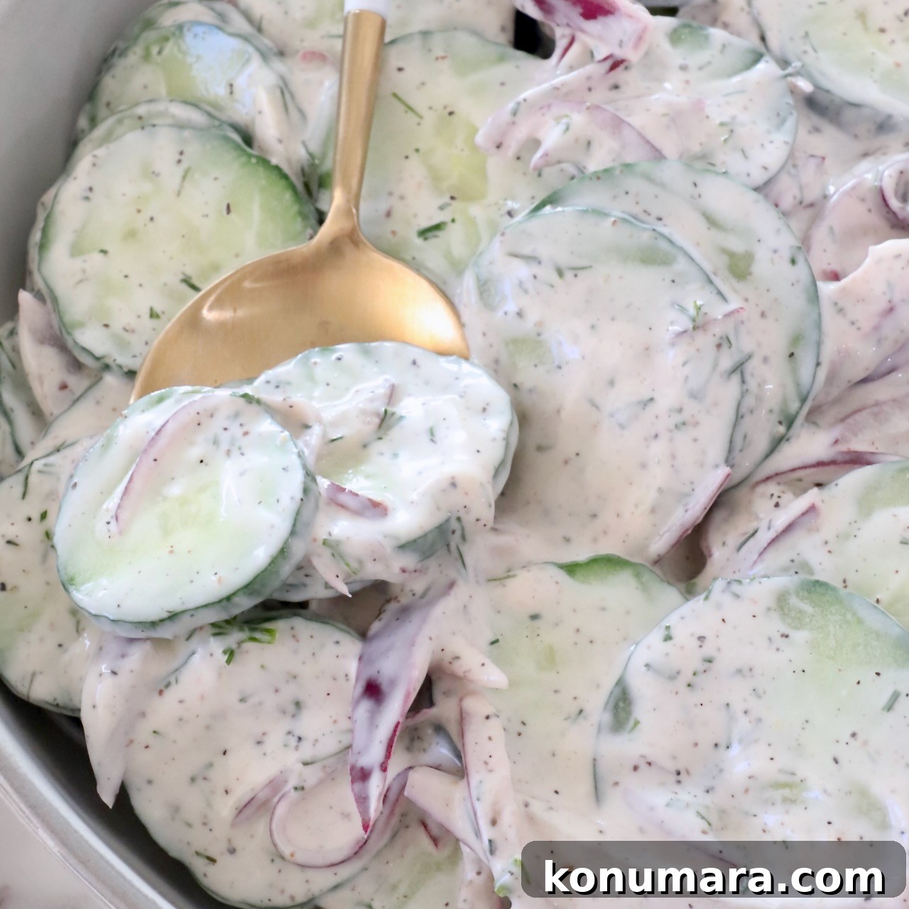 sliced cucumbers and red onions tossed in a creamy dressing in a bowl with a gold spoon