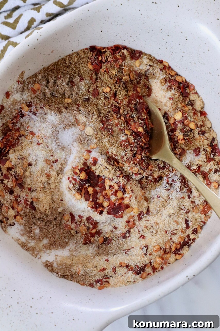 Homemade Thai Seasoning Blend: Authentic Recipe 2 A white bowl contains a mix of red chili flakes, brown spices, and white salt, stirred with a golden spoon. A patterned cloth is partially visible.