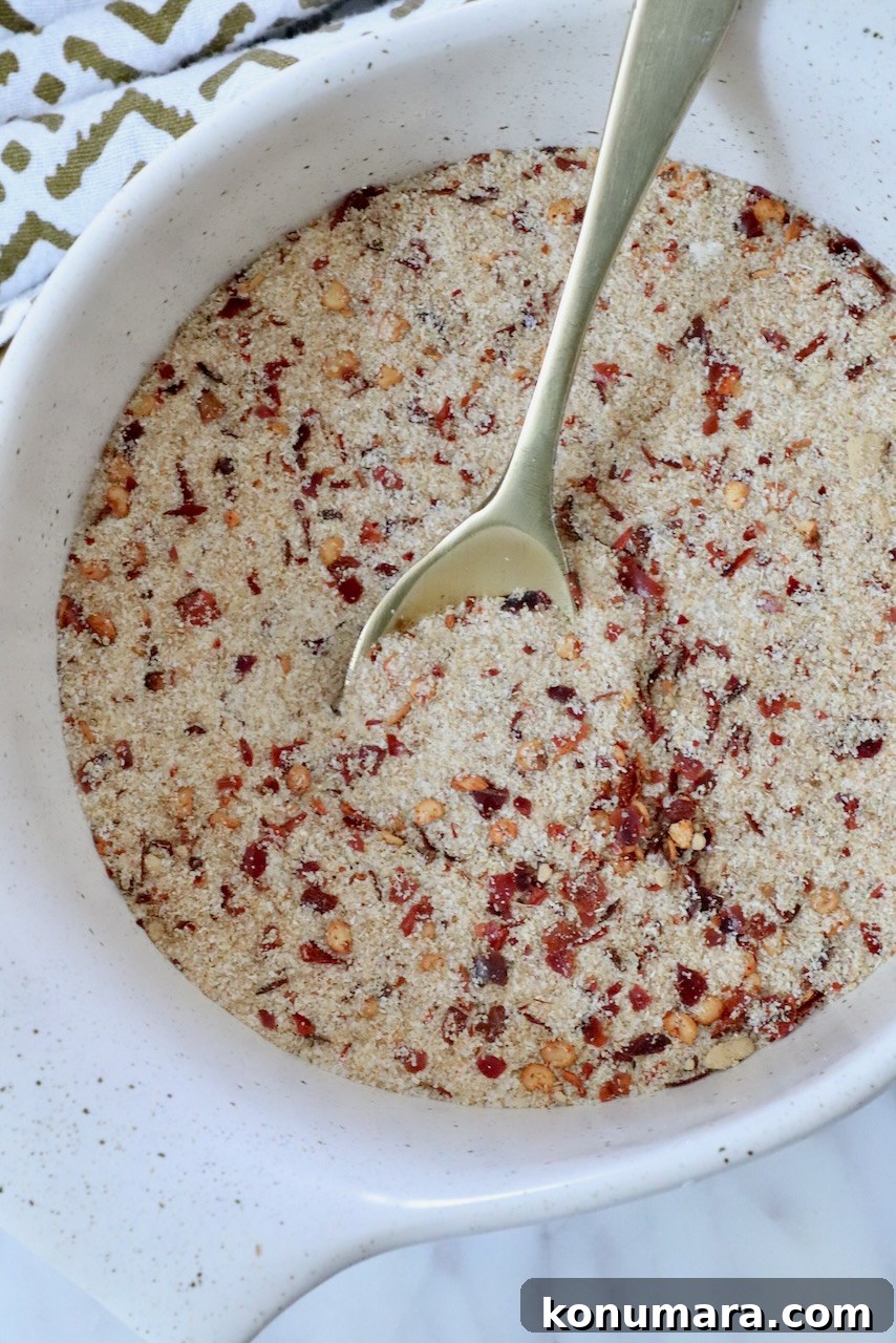 Homemade Thai Seasoning Blend: Authentic Recipe 4 A white bowl filled with a dry mixture of salt, spices and red pepper flakes, featuring a metal spoon. A patterned cloth is partially visible beside the bowl.