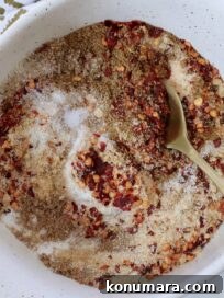 A white bowl contains a mix of red chili flakes, brown spices, and white salt, stirred with a golden spoon. A patterned cloth is partially visible.