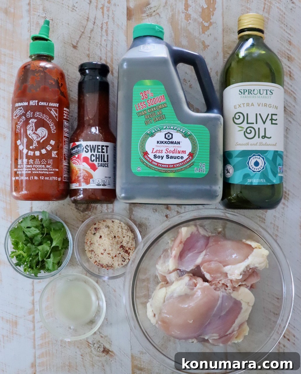 Authentic Thai Basil Chicken Stir-Fry (Pad Kra Pao) 3 Ingredients for a recipe are arranged on a wooden surface. From left to right: Sriracha, sweet chili sauce, soy sauce, olive oil, cilantro, thai seasoning, lime juice, and raw chicken thighs.