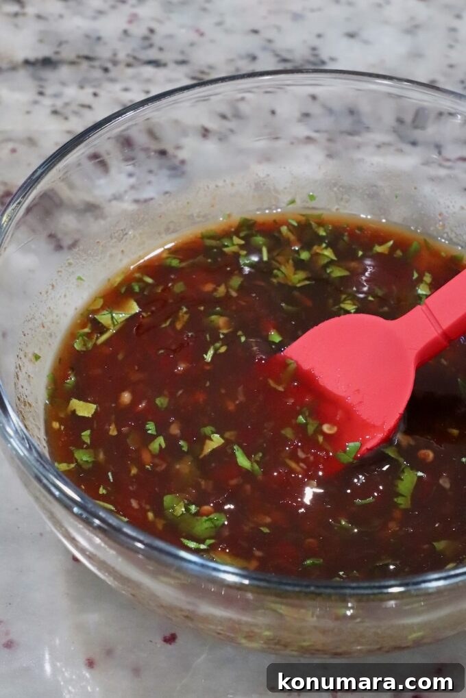 Authentic Thai Basil Chicken Stir-Fry (Pad Kra Pao) 5 A glass bowl filled with a reddish-brown sauce, featuring chopped herbs, is on a marble surface. A red spatula rests in the bowl.