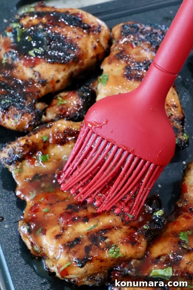 Authentic Thai Basil Chicken Stir-Fry (Pad Kra Pao) 7 Grilled chicken thighs being brushed with glossy sauce using a red brush, showing charred marks and fresh herbs on a dark surface.