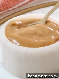 A small white ceramic bowl filled with smooth peanut butter dressing with a spoon dipping in.