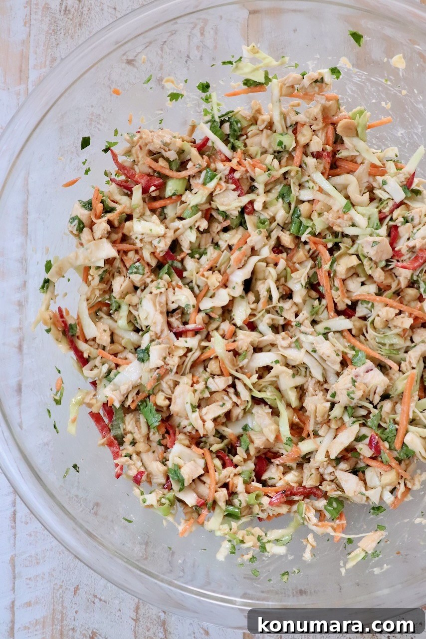 A glass bowl filled with colorful chicken salad on a light wooden surface. The salad includes shredded cabbage, carrots, red peppers, and green herbs, giving a fresh, vibrant look.