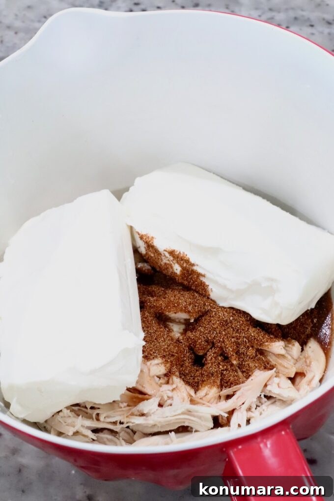 Spicy Nashville Hot Chicken Dip Recipe 4 A red mixing bowl with shredded chicken, two blocks of cream cheese, and a sprinkle of spices.