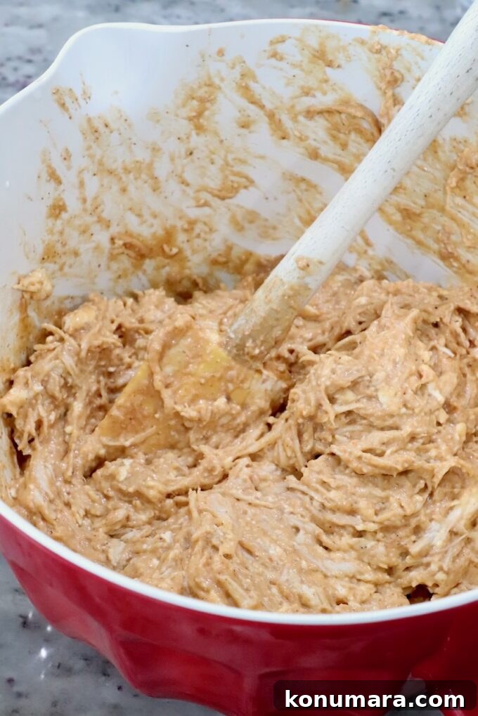 Spicy Nashville Hot Chicken Dip Recipe 5 A red bowl filled with a thick, creamy chicken and cheese mixture being mixed with a wooden spoon.