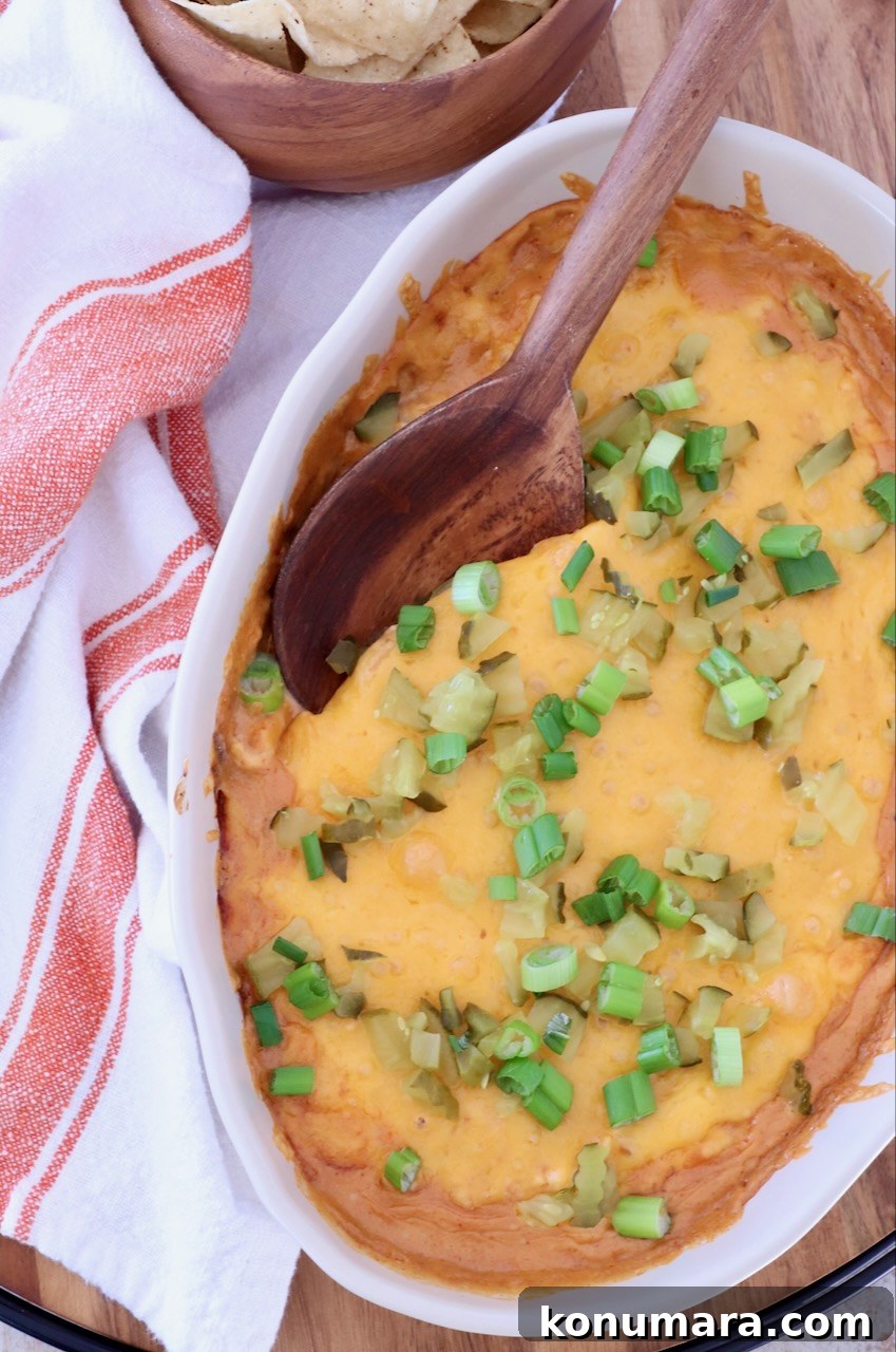 Spicy Nashville Hot Chicken Dip Recipe 8 Close-up of a baked cheesy dip in an oval dish topped with green onions and pickles. A wooden spoon rests on top. Tortilla chips in a bowl and a striped towel are nearby.
