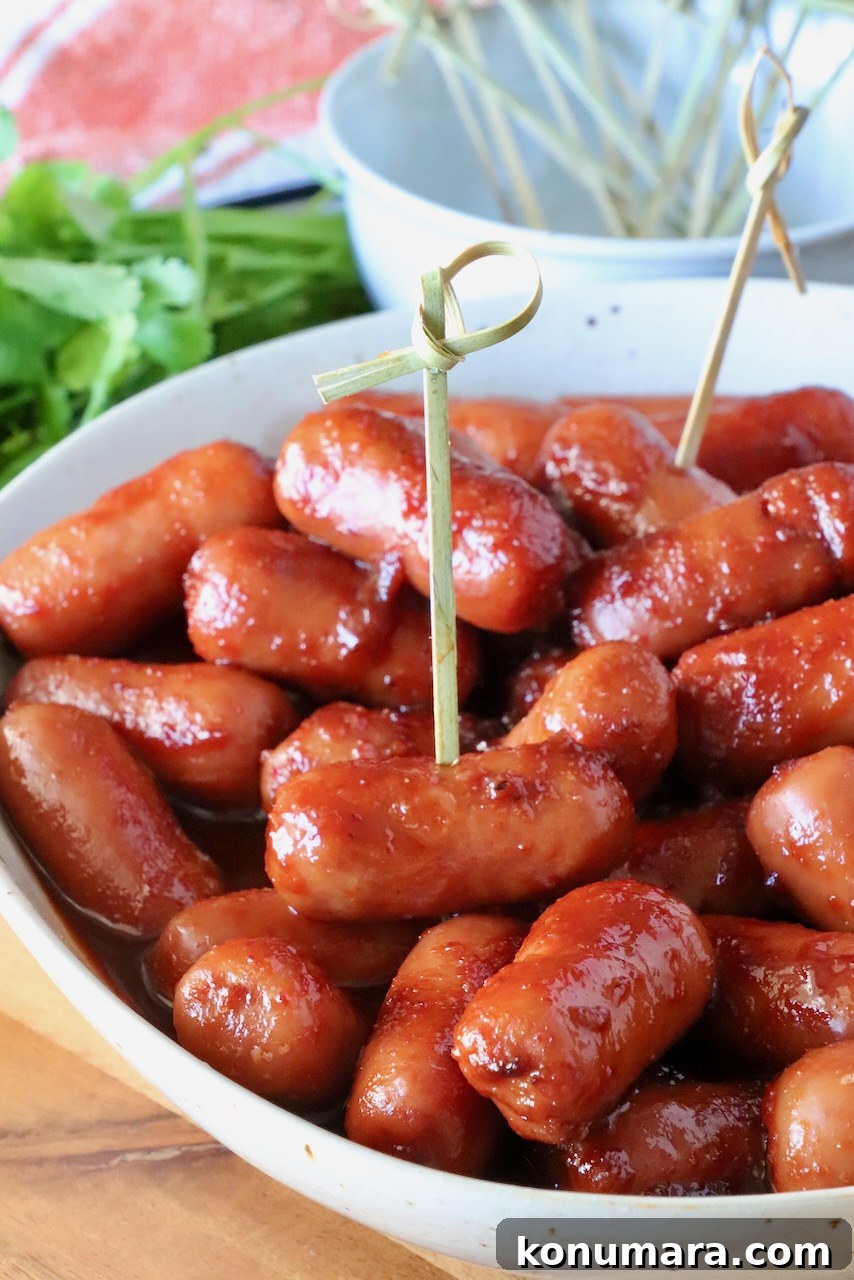 Chipotle Honey Glazed Little Smokies 2 A close-up of glazed cocktail sausages in a white bowl with toothpicks, placed on a wooden table. Fresh cilantro is visible in the background.
