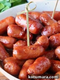 A close-up of glazed cocktail sausages in a white bowl with toothpicks, placed on a wooden table. Fresh cilantro is visible in the background.