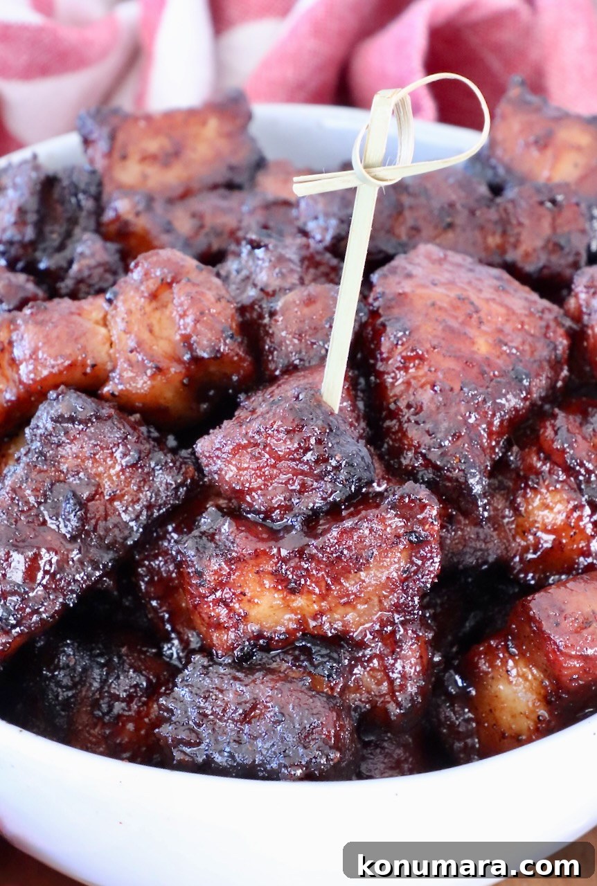 Close-up of glazed, caramelized pork belly burnt ends in a white bowl with a skewer. The meat looks juicy and tender.