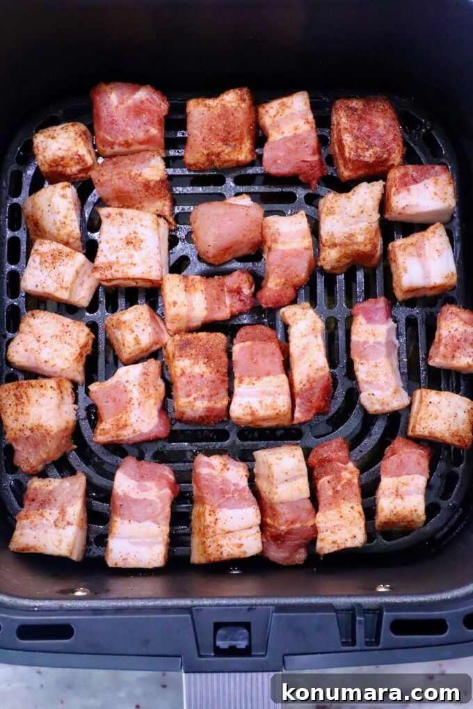 Cubed pork belly pieces, seasoned with spices, are evenly spread in the basket of an air fryer. The meat is pink and golden, ready for cooking.