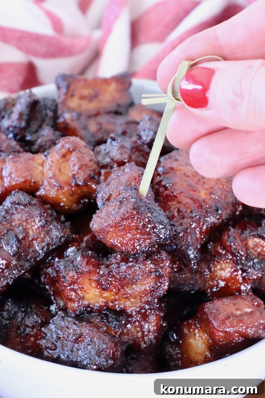 A hand with red nail polish holds a toothpick, picking up a glazed, caramelized pork belly bite from a bowl filled with similar pieces. Red-striped cloth is in the background.