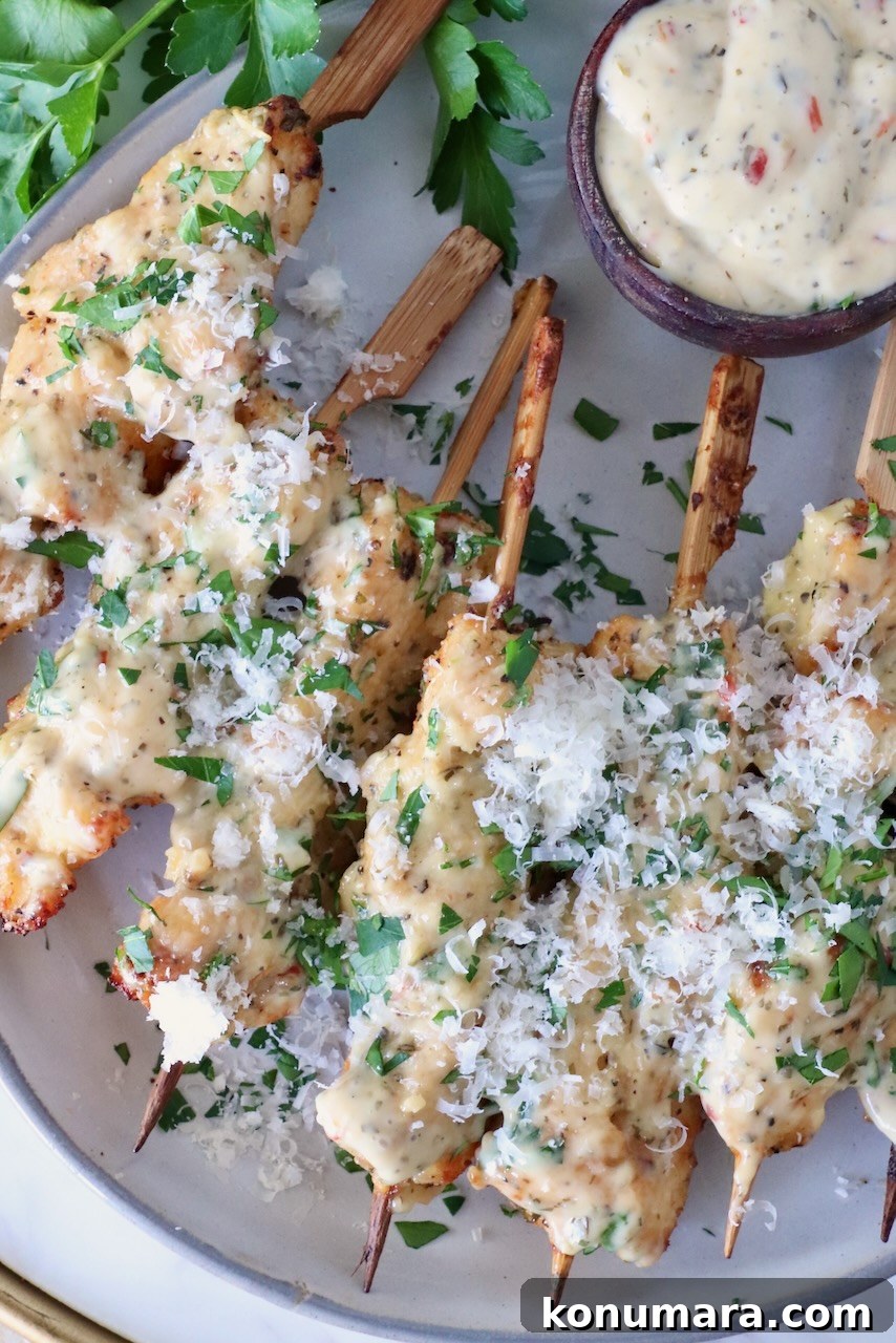 cooked chicken skewers topped with creamy sauce and shredded cheese, garnished with chopped parsley, served beside a small bowl of dipping sauce.