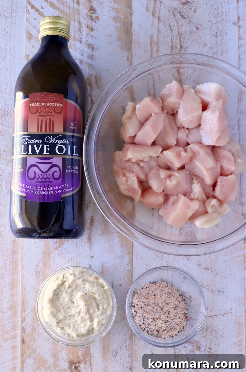 Bottle of olive oil next to a bowl of raw chicken chunks, with small bowls of creamy sauce and seasoning on a rustic wooden surface.