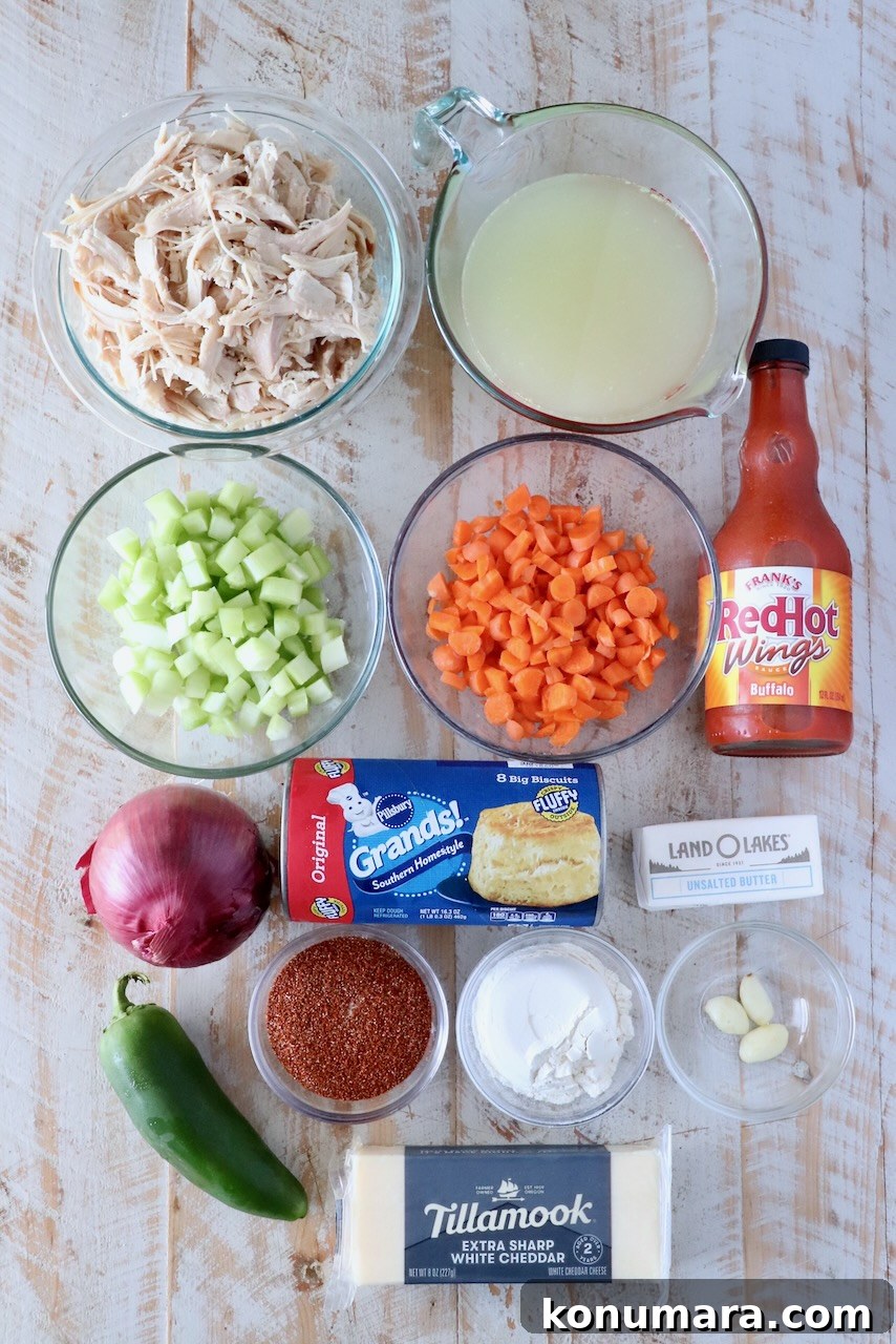 Overhead view of cooking ingredients: shredded chicken, broth, diced celery, carrots, hot sauce, onion, jalapeño, biscuits, butter, seasonings, and cheddar cheese