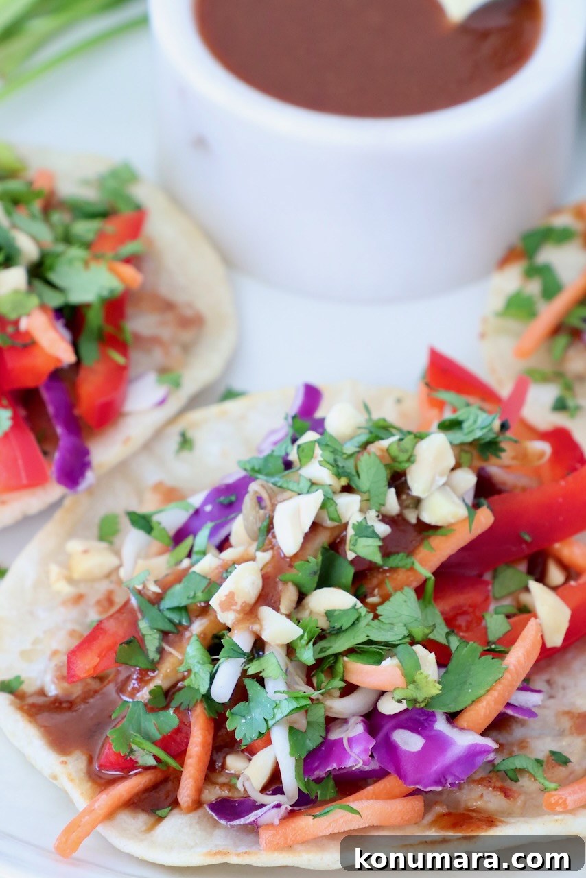 Close-up of pad thai tacos with vibrant toppings like red peppers, carrots, purple cabbage, and fresh cilantro, next to a bowl of sauce.