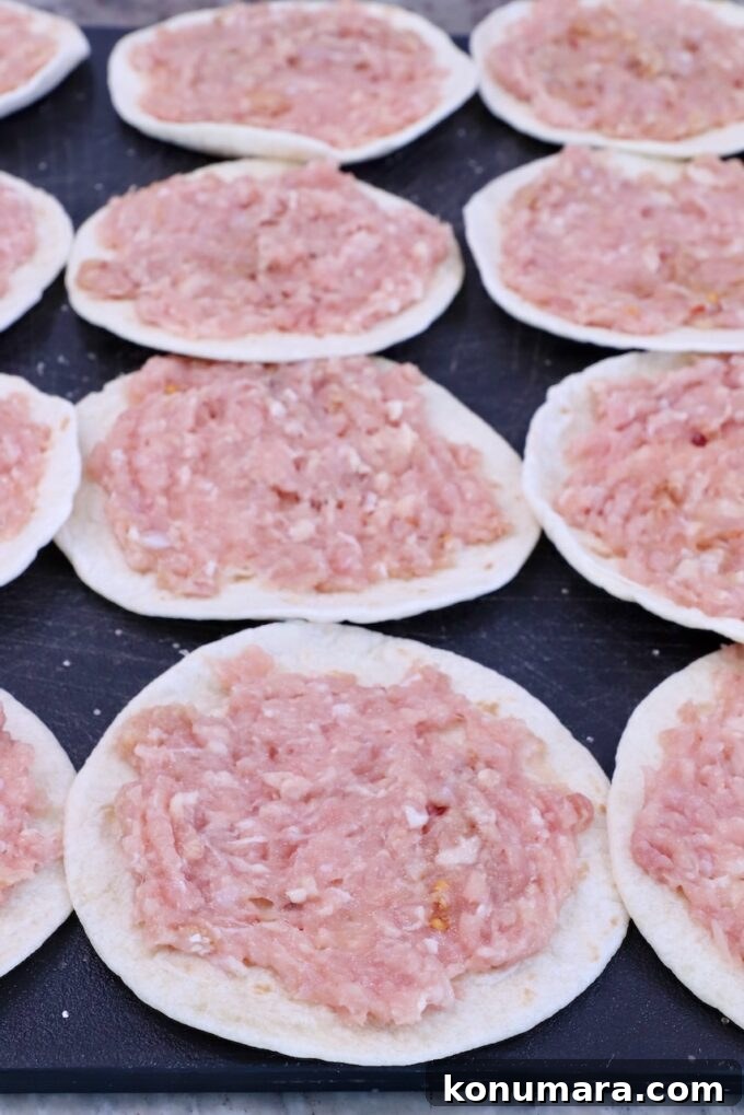 Raw ground chicken is spread on small, round tortillas laid out on a dark surface.