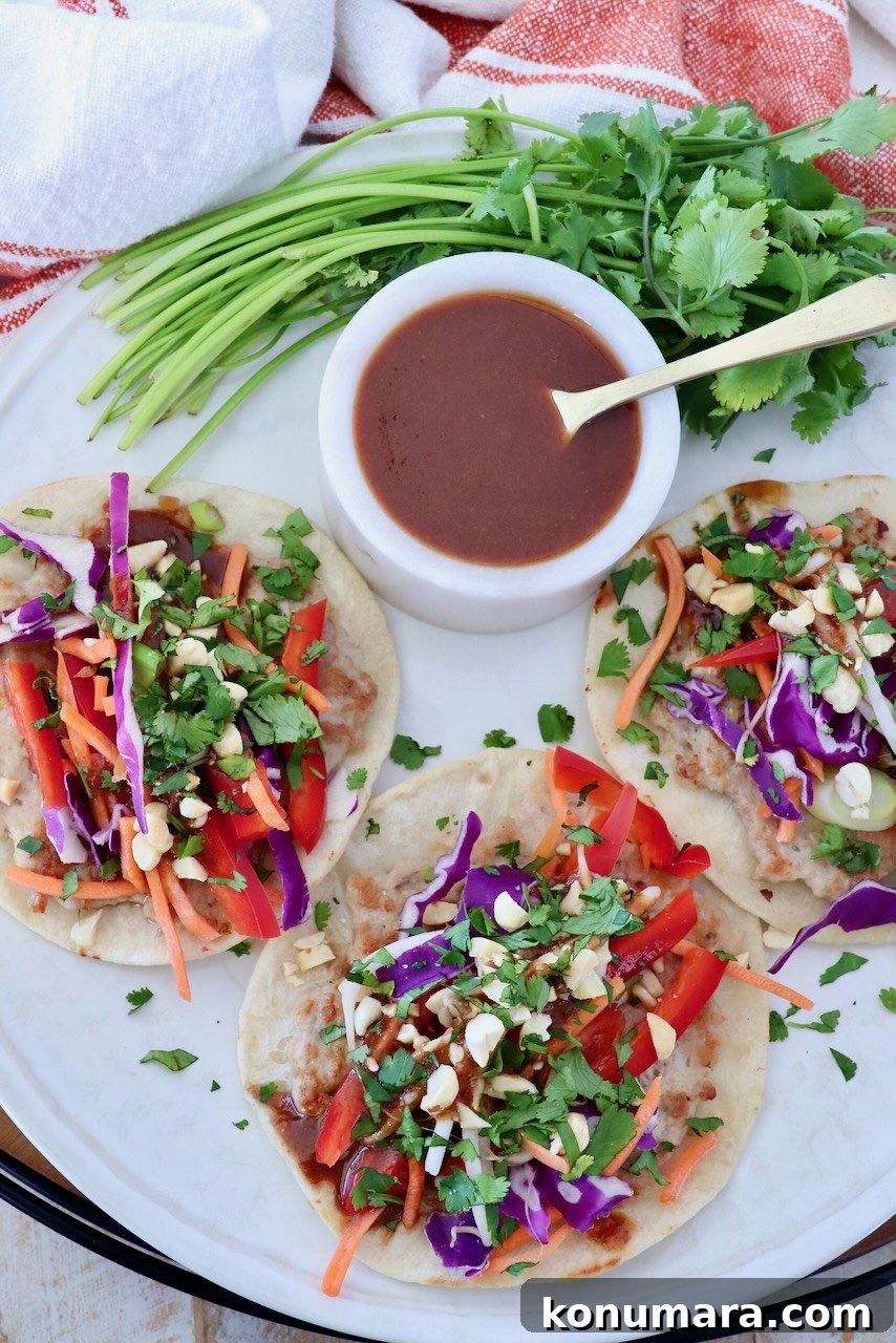 Plate of colorful pad thai tacos topped with vibrant red peppers, purple cabbage, carrots, and fresh cilantro. A ramekin of sauce and cilantro garnish beside.