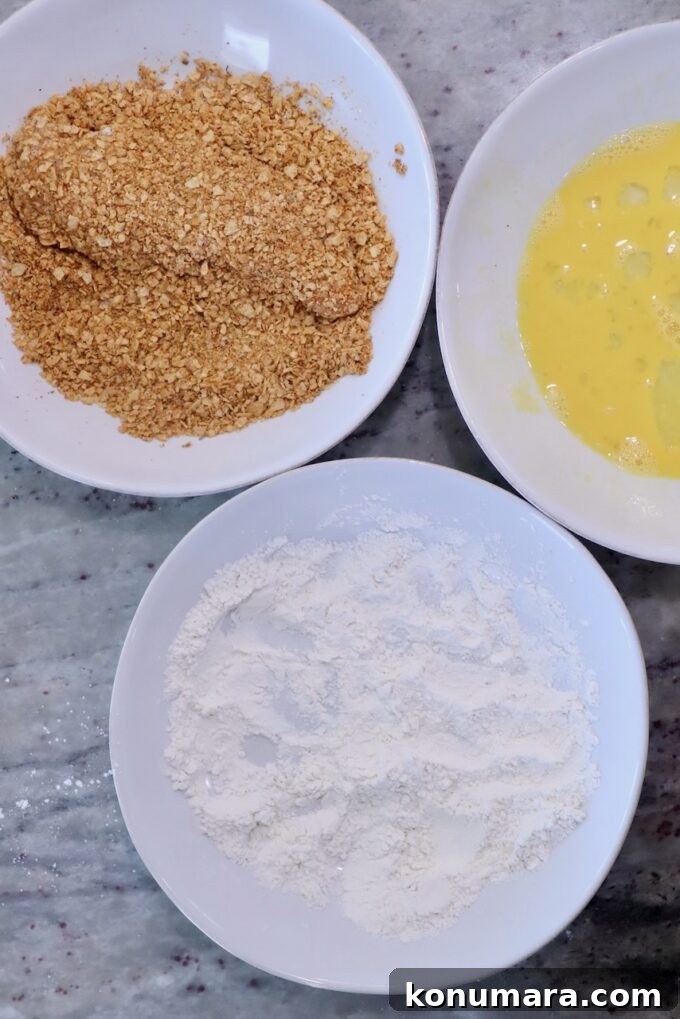 Crispy Mexican-Style Chicken Parmesan with Chipotle Sauce 4 Three bowls on a marble surface contain ingredients for breading: flour, beaten eggs, and tortilla chip crumbs. The setup is ready for coating food.