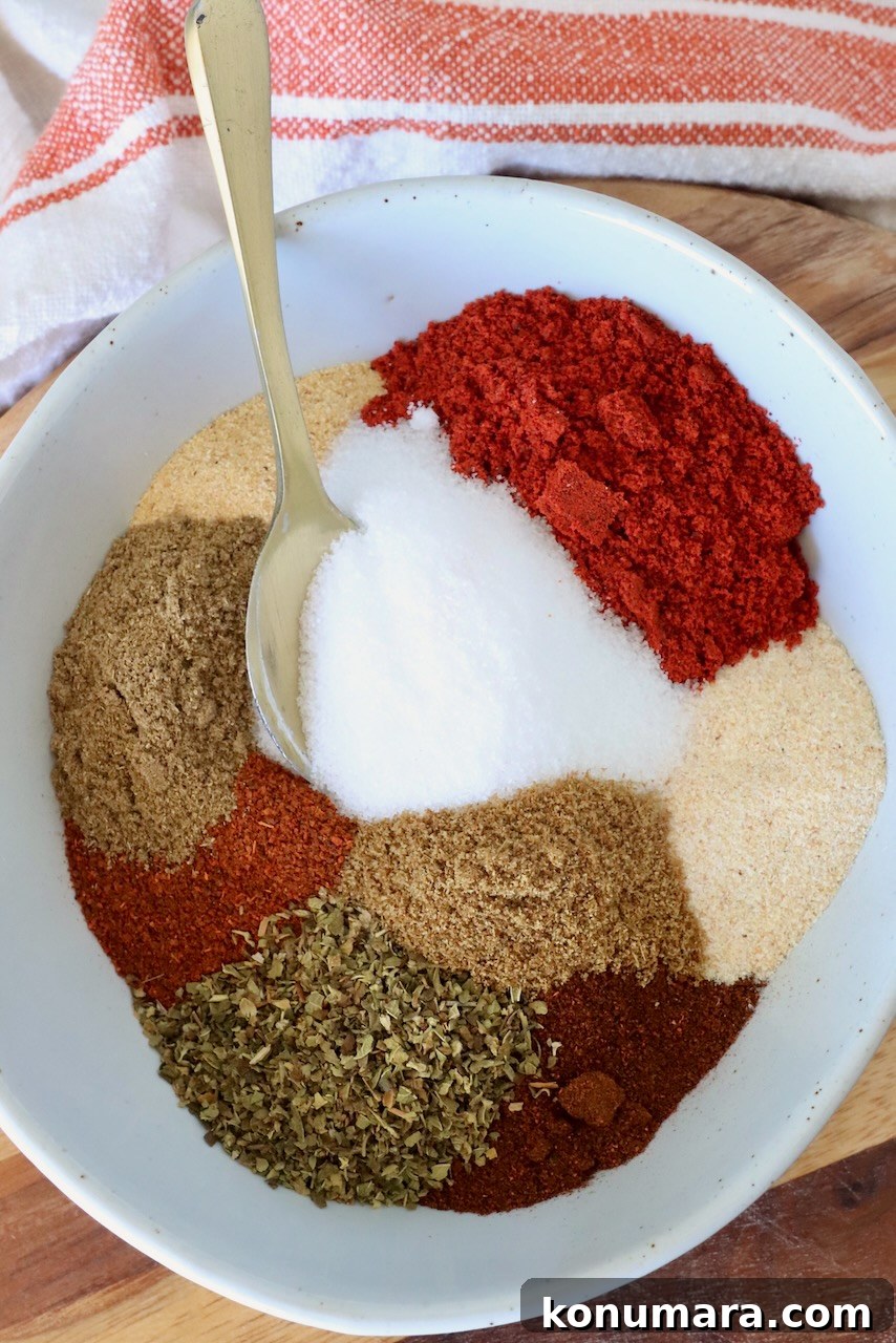 A white bowl filled with colorful spices, including red chili powder, white salt, brown cumin, and green oregano. A gold spoon rests on the side.