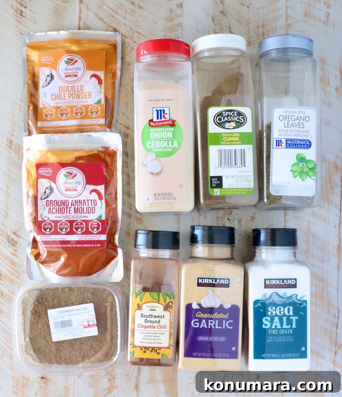 A variety of seasoning packages are arranged on a wooden surface, featuring chili powders, granulated onion, cumin, oregano, garlic, sea salt, and more.