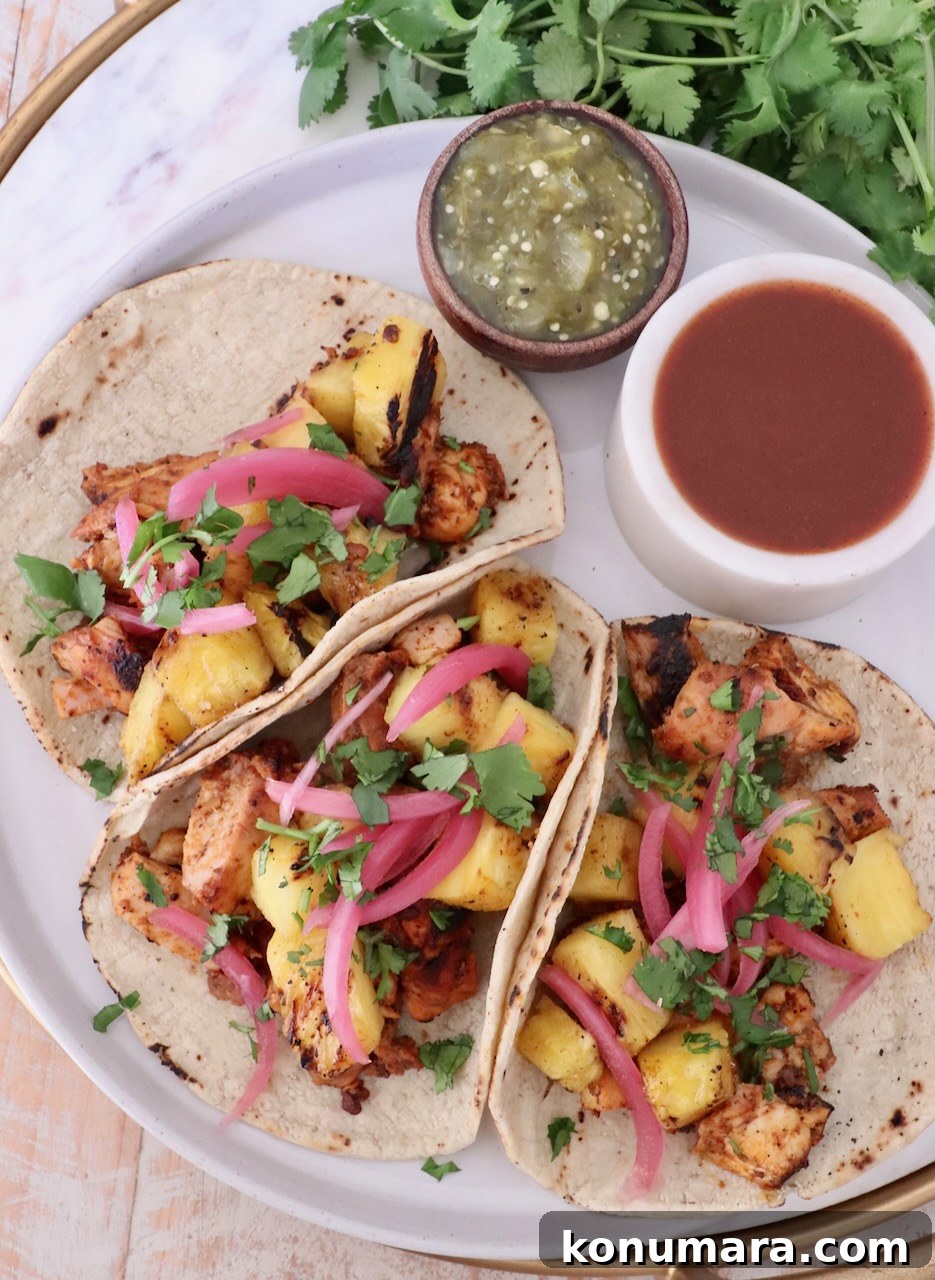 How to Make Chicken Al Pastor Tacos with Pineapple 6 Three tacos filled with grilled chicken, pineapple, pickled onions, and cilantro on a plate. Beside them are bowls of green salsa and hot sauce.