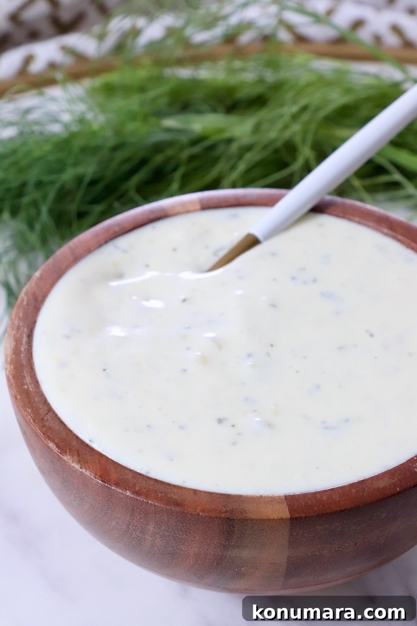 Creamy Dill Pickle Ranch Dressing Recipe 2 A wooden bowl filled with creamy, pickle ranch dressing, speckled with herbs. A white spoon rests inside. Fresh dill fronds are in the background.