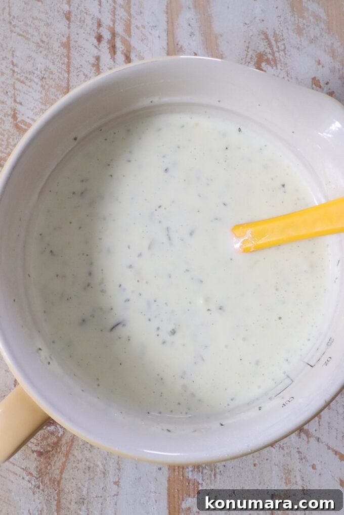 Creamy Dill Pickle Ranch Dressing Recipe 5 Creamy ranch dressing with herbs in a yellow mixing bowl, with a yellow spatula resting inside.