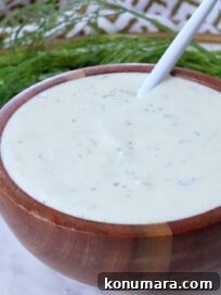 A wooden bowl filled with creamy, pickle ranch dressing, speckled with herbs. A white spoon rests inside. Fresh dill fronds are in the background.