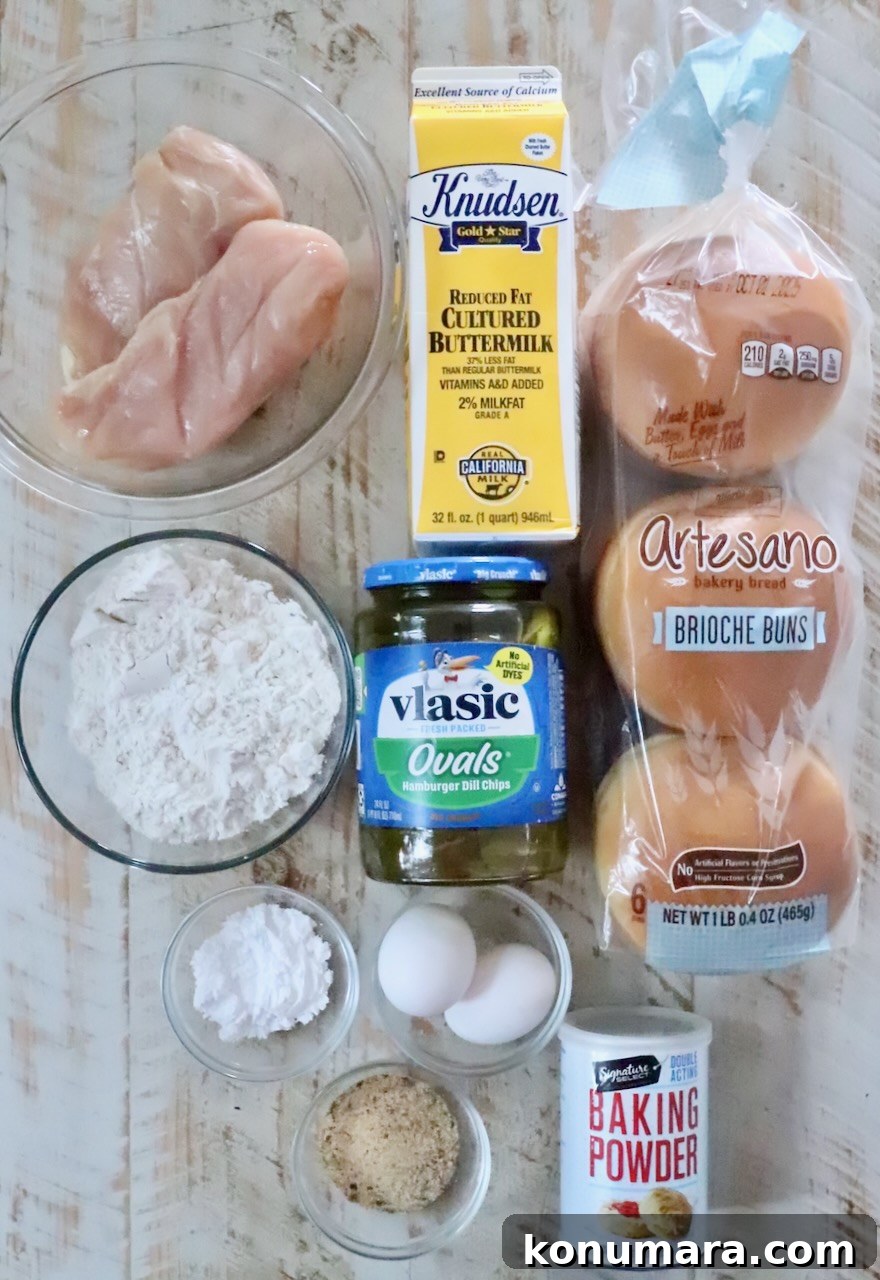 Crispy Air Fryer Chicken Sandwich Recipe 3 Ingredients for a chicken sandwich are neatly arranged: raw chicken, buttermilk, brioche buns, flour, pickles, eggs, baking powder, and chicken seasoning.