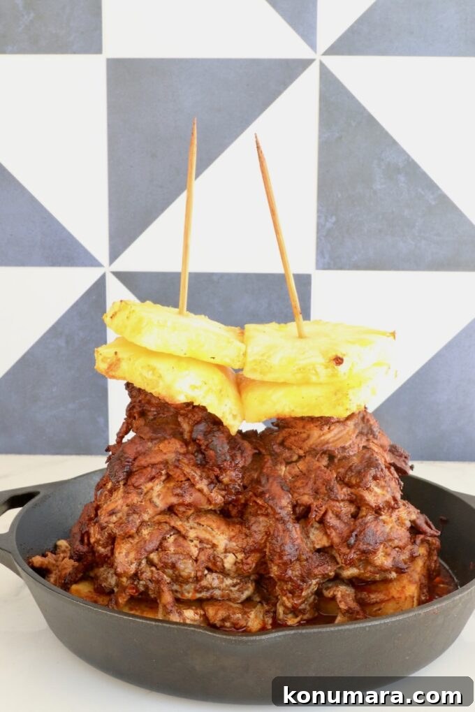 Slow-Roasted Al Pastor Pork Tacos 6 A stack of marinated, cooked pork layered on skewers, topped with pineapple slices, in a cast iron skillet.