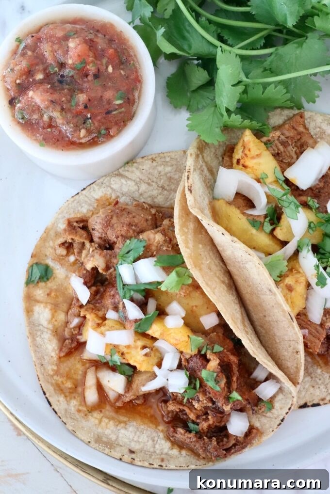 Slow-Roasted Al Pastor Pork Tacos 7 Two tacos with pork al pastor, pineapple, onion, and cilantro on tortillas, accompanied by a bowl of salsa and fresh cilantro, arranged on a plate.