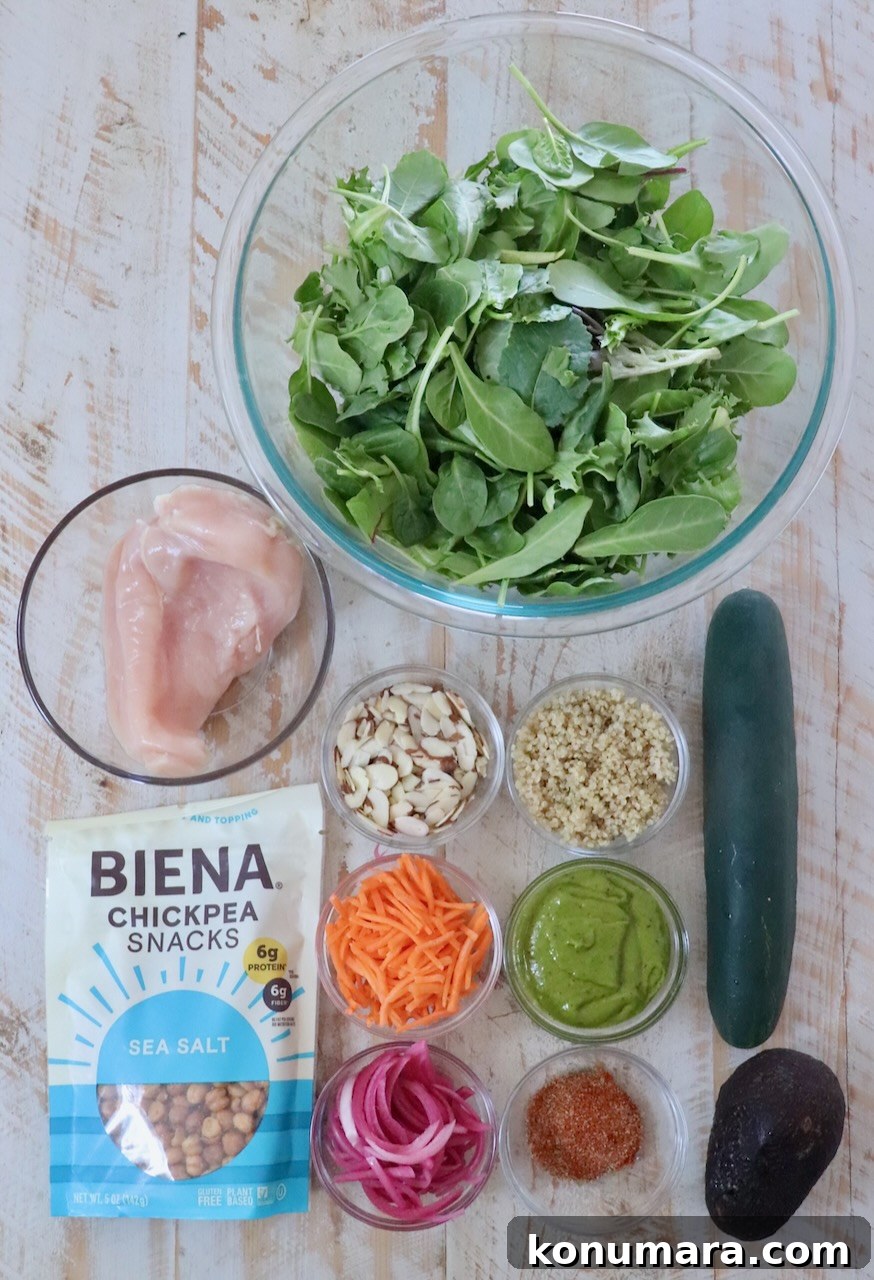 Top view of salad ingredients on a wooden surface: mixed greens, chicken, cucumber, avocado, quinoa, sliced almonds, chickpea snacks, shredded carrots, and pickled onions.