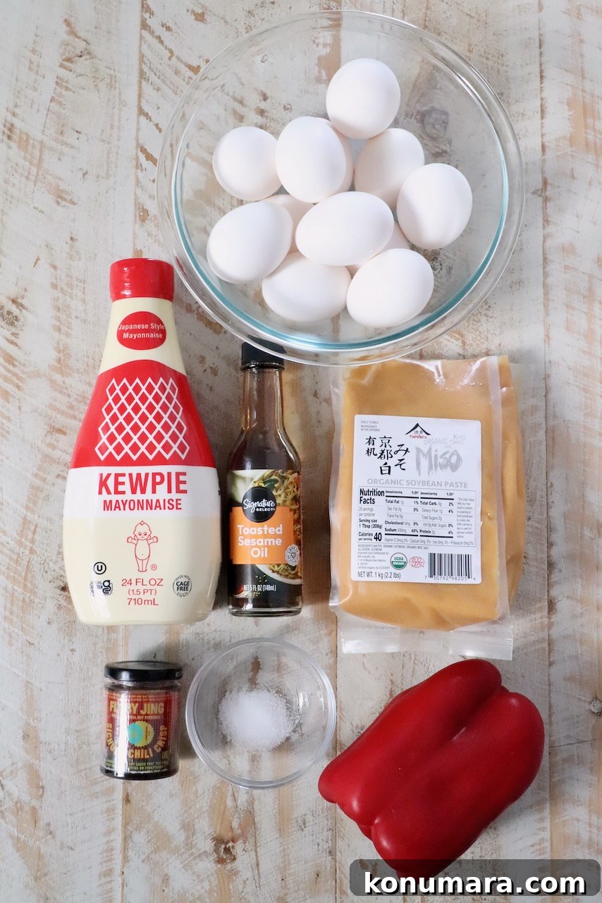 Spicy Miso Egg Salad with Chili Crisp Recipe 5 A flat lay of cooking ingredients on a wooden surface includes a bowl of eggs, Kewpie mayonnaise, toasted sesame oil, miso, a jar of chili paste, salt, and a red bell pepper.
