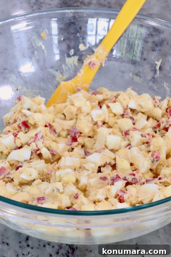 Spicy Miso Egg Salad with Chili Crisp Recipe 9 A clear glass bowl on a marble countertop holds a miso egg salad with minced red peppers. A yellow spatula rests in the bowl.
